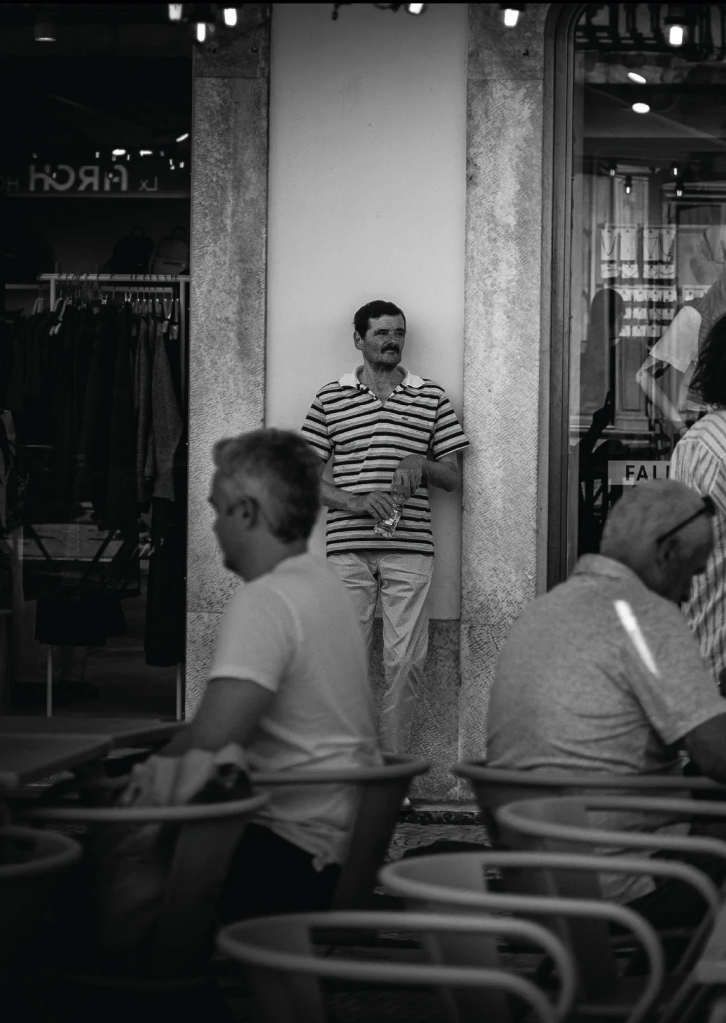 Um homem de camisa às riscas à porta de um café, entre outras pessoas na rua.